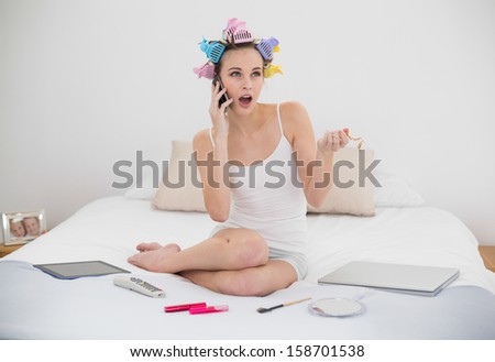 Shocked natural brown haired woman in hair curlers calling with her mobile phone in bright bedroom