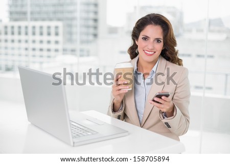Cheerful businesswoman holding disposable cup and smartphone in bright office