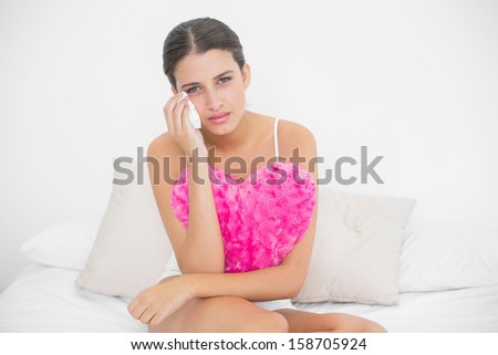Stern young brown haired model in white pajamas wiping her tears in bright bedroom