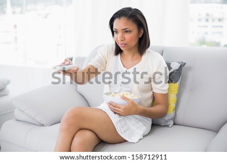 Serious young dark haired woman in white clothes changing tv chanel while eating popcorn in a living room