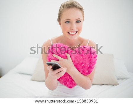 Natural cheerful blonde holding heart pillow and smartphone in bright bedroom