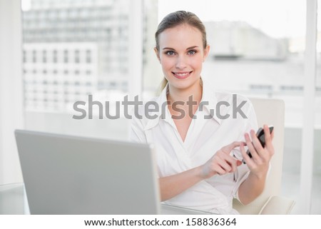 Smiling businesswoman using laptop and texting on smartphone in her office
