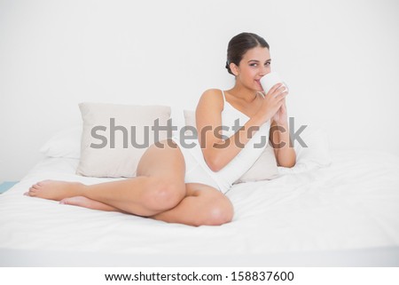 Calm young brown haired model in white pajamas drinking coffee in bright bedroom
