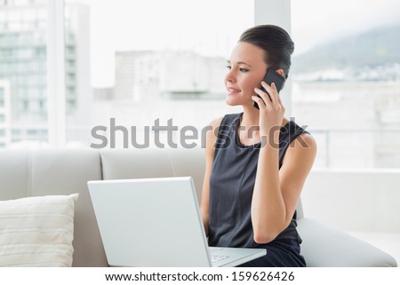 Beautiful well dressed young woman using laptop and cellphone on sofa at bright home