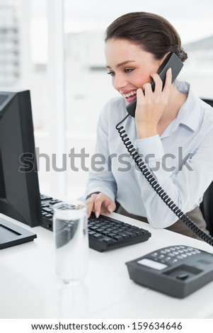 Smiling elegant businesswoman using land line phone and computer in a bright office