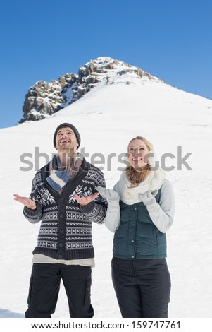 Cheerful couple in warm clothing with hands open looking up in front of snowed hill and clear blue sky