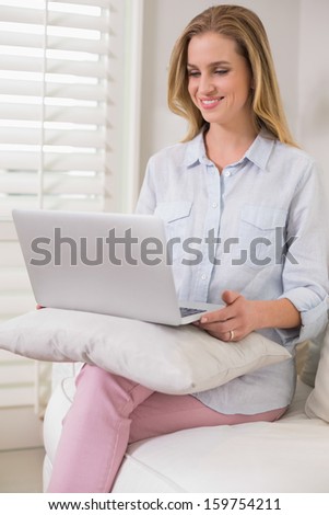 Happy casual blonde sitting on couch using laptop in bright living room