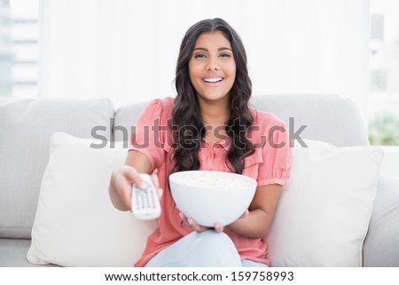 Happy cute brunette sitting on couch holding popcorn bowl in bright living room