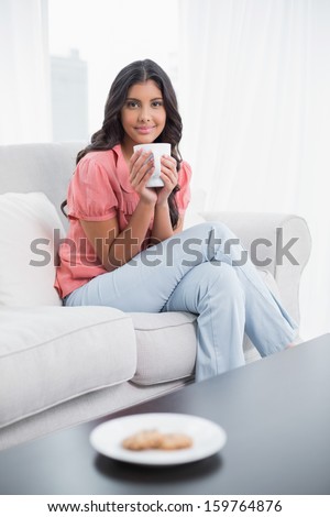 Content cute brunette sitting on couch holding cup of coffee in bright living room
