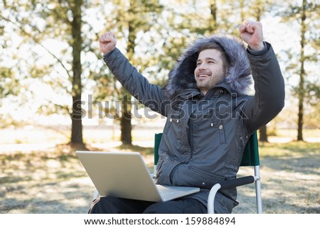 Cheerful young man in fur hood jacket with laptop clenching fists in the forest on a winter day