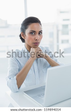 Serious dark haired woman looking at camera sitting at her desk in bright office