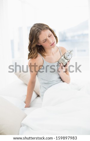 Annoyed slender woman sitting on her bed under the cover holding the alarm clock