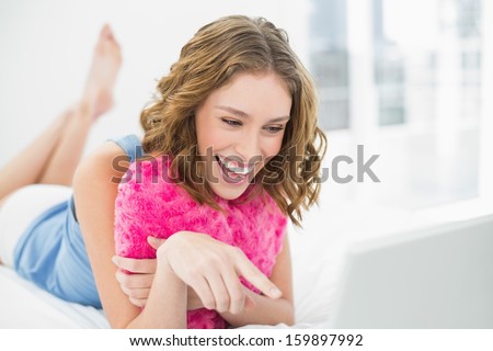 Lovely laughing woman cuddling with a heart pillow pointing at her notebook lying on her bed