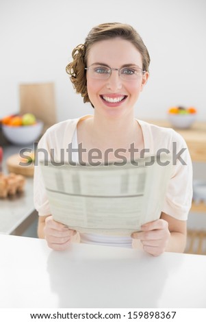 Cute woman holding newspaper sitting in her kitchen at home wearing glasses