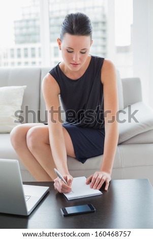Beautiful young businesswoman writing notes by laptop on sofa at a bright home