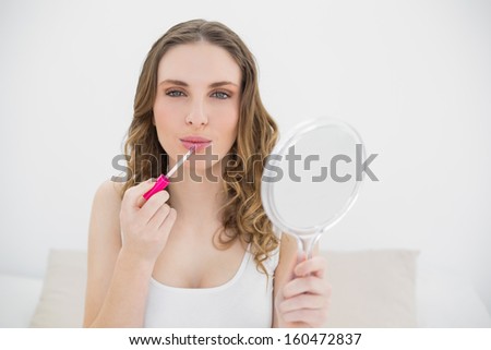 Young woman using lip gloss and looking into the camera while holding a mirror