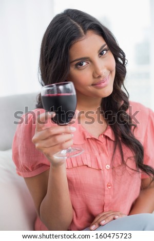 Smiling cute brunette sitting on couch holding wine glass in bright living room
