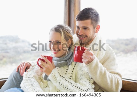 Young couple in winter clothing drinking coffee against cabin window