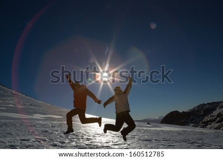 Full length of a silhouette couple jumping on snow covered landscape against the sun and clear blue sky