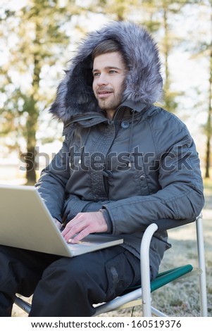 Young man in fur hood jacket using laptop in the forest on a winter day