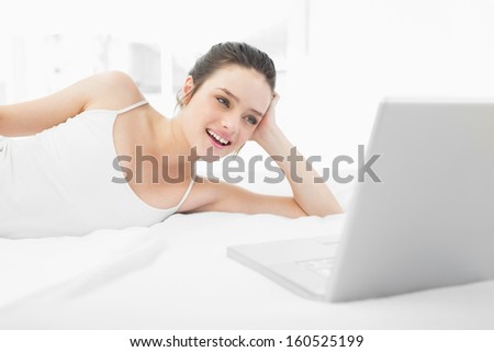 Smiling casual young woman looking at laptop while lying in bed