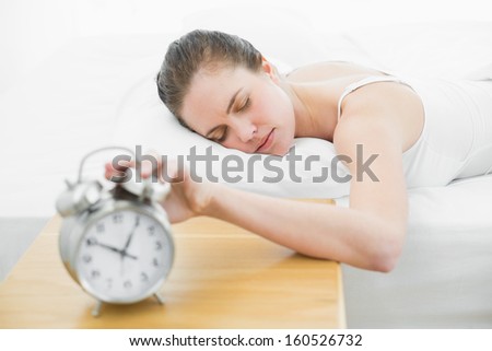 Sleepy woman in bed extending hand to blurred alarm clock at bedroom