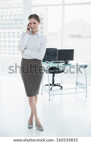 Gleeful calm businesswoman phoning with her smartphone standing in her office smiling at camera