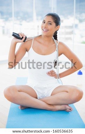 Smiling toned brunette sitting on floor with skipping rope in bright room