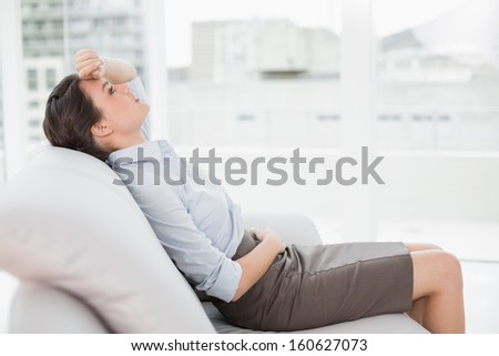 Side view of a tired beautiful well dressed young woman sitting on sofa at bright home