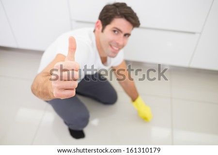 Portrait of a smiling young man cleaning the kitchen floor while gesturing thumbs up at house