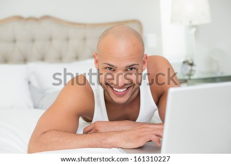 Portrait of a smiling casual bald young man using laptop in bed at home
