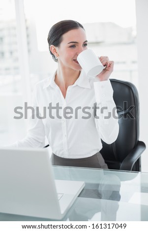 Smiling businesswoman drinking coffee while using laptop at desk in a bright office