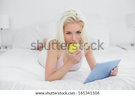 Portrait of a casual young blond with tablet PC eating an apple in bed at home