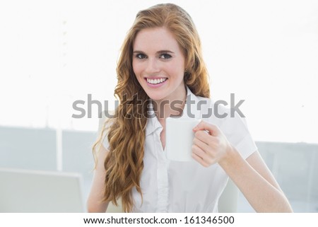 Portrait of a smiling businesswoman drinking coffee using laptop at desk in a bright office