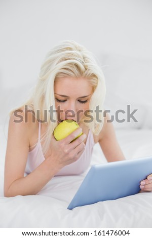 Casual young blond eating an apple while using tablet PC in bed at home