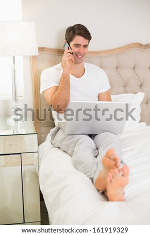 Casual smiling young man using cellphone and laptop in bed at home