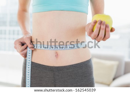 Close up mid section of a young woman measuring waist as she holds apple in fitness studio