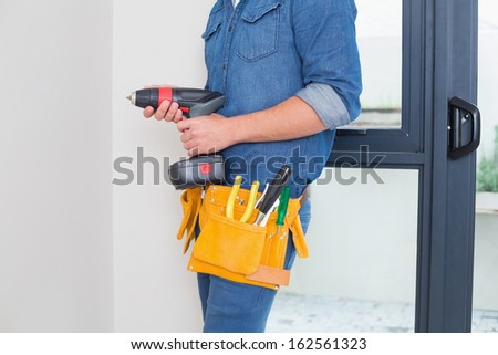 Mid section of a handyman with drill and tool belt by the wall