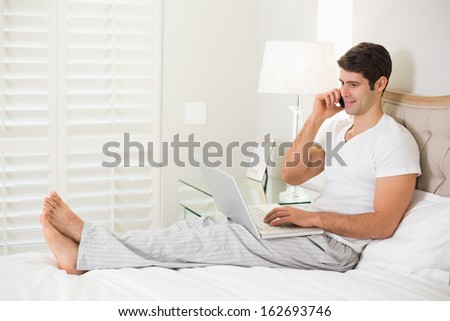 Side view of a casual smiling young man using cellphone and laptop in bed at home