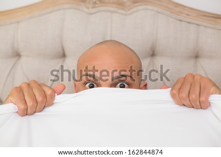 Close up portrait of a young shocked bald man covering face with sheet in bed at home