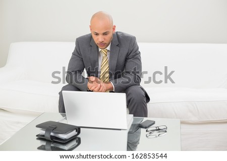 Serious elegant young businessman looking at laptop with diary on table at home