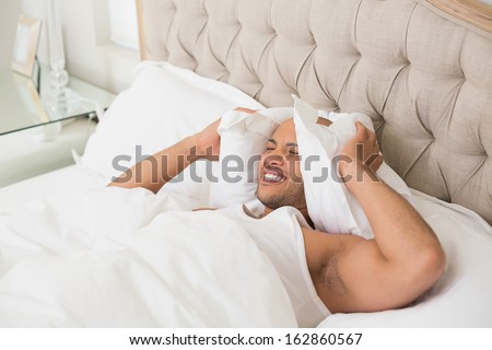 Young sleepy man covering ears with pillow in bed at home