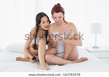 Full length of two shocked young female friends text messaging on bed at home
