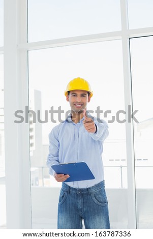 Portrait of a smiling young architect in yellow hard hat with clipboard gesturing thumbs up in a bright office