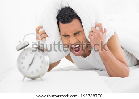 Angry young man covering ears with pillow and shouting with alarm clock in foreground at bed