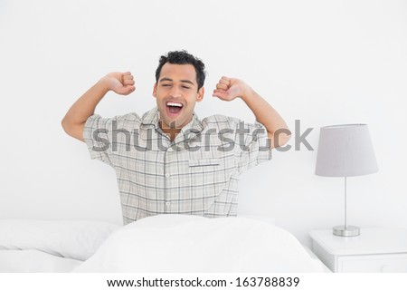 Smiling young man waking up in bed and stretching his arms