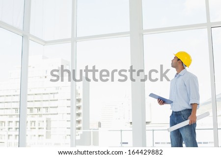 Side view of a handyman in yellow hard hat with clipboard and blueprint looking up at window in a bright office