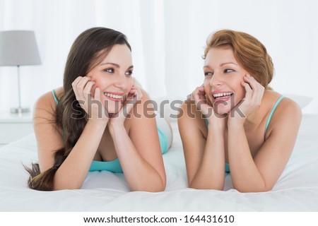 Two cheerful young female friends in teal tank tops lying in bed at home