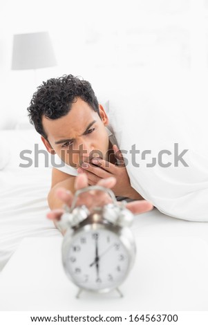 Close-up of a sleepy young man in bed extending hand to alarm clock at home