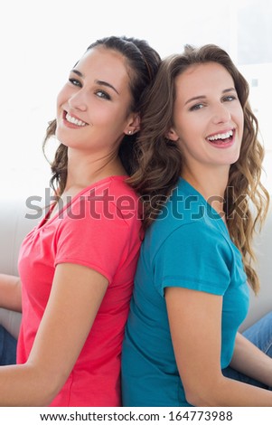 Side view portrait of two smiling young female friends sitting back to back in the living room at home